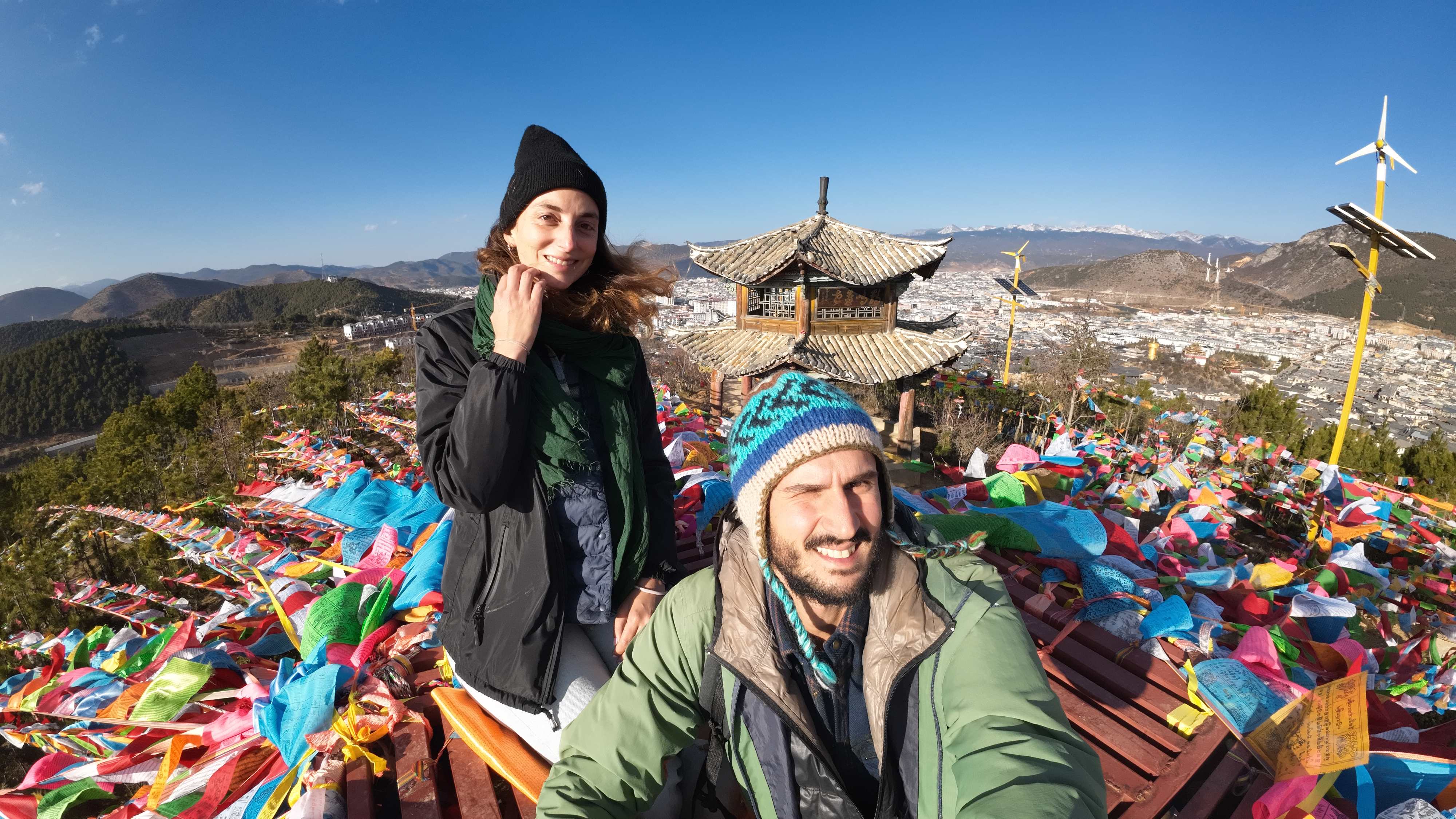 Sofi & Lele smiling above Tibetan Flags and view of Shangri La from a hill